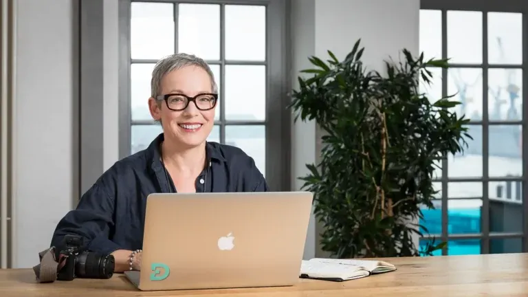 Adobe Mitarbeiterin Bettina Reinemann sitzt lächelnd vor ihrem MacBook mit Kamera und Notizbuch in einem hellen Loftbüro mit Hafenblick, fotografiert von Frank Erpinar