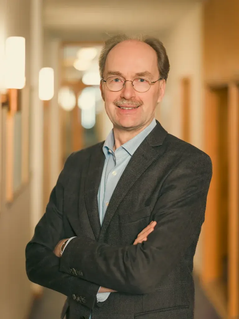 Martin Oldeland steht selbstbewusst mit verschränkten Armen im dunklen Sakko im Büroflur von B.A.U.M. e.V.