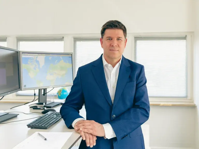 Seriöser Mann im blauen Anzug lehnt an einem Schreibtisch vor zwei Monitoren mit Weltkarte in einem modernen Büro