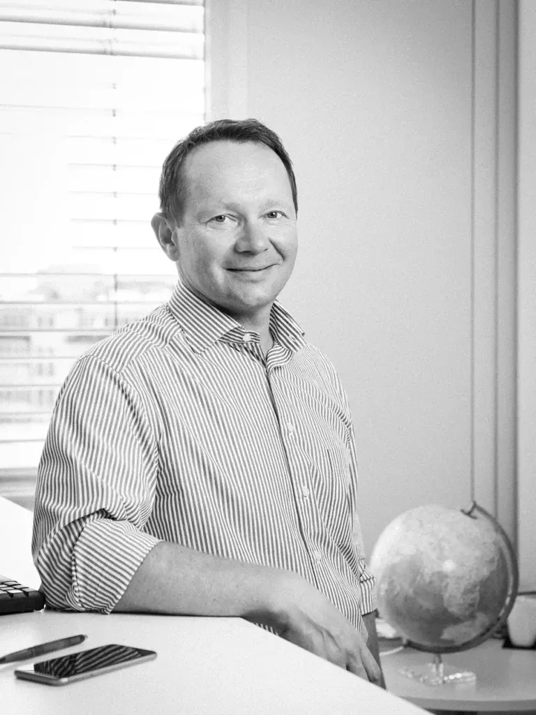 Schwarz-Weiß-Portrait eines Mitarbeiters der Skuld Germany GmbH am Schreibtisch mit kleinem Globus und freundlichem Lächeln, fotografiert von Frank Erpinar