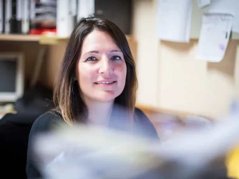 Dunkelhaarige Frau am Arbeitsplatz lächelt freundlich in die Kamera, umgeben von Büromaterial und Ordnern, fotografiert von Frank Erpinar